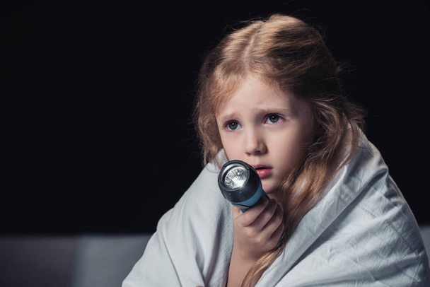 scared kid sitting under blanket and holding flashlight isolated on black - Photo, Image