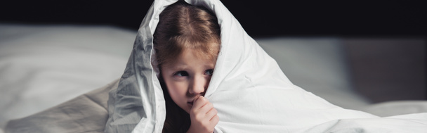 Panorama-Aufnahme eines verängstigten Kindes, das sich unter einer Decke versteckt, isoliert auf schwarz - Foto, Bild