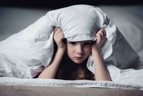 frightened kid hiding under blanket and looking at camera isolated on black - Foto, Imagem