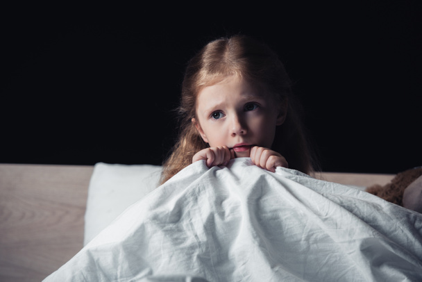 scared child looking away while sitting on bedding in dark bedroom isolated on black - Photo, Image