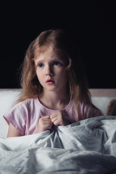 przestraszony dzieciak patrząc z dala siedząc na łóżku izolowane na czarnym - Zdjęcie, obraz