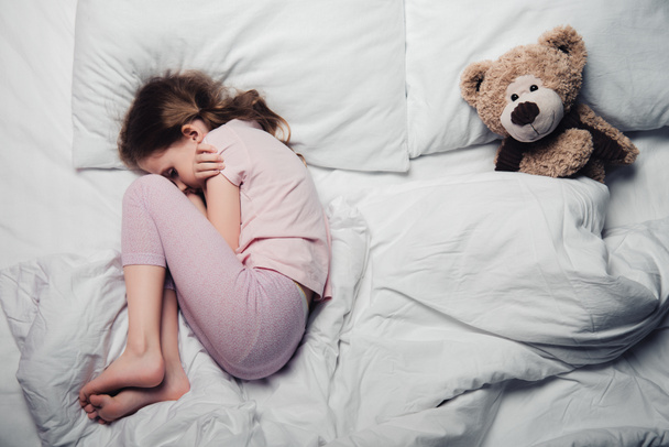 top view of scared child lying on white bedding near teddy bear - Φωτογραφία, εικόνα