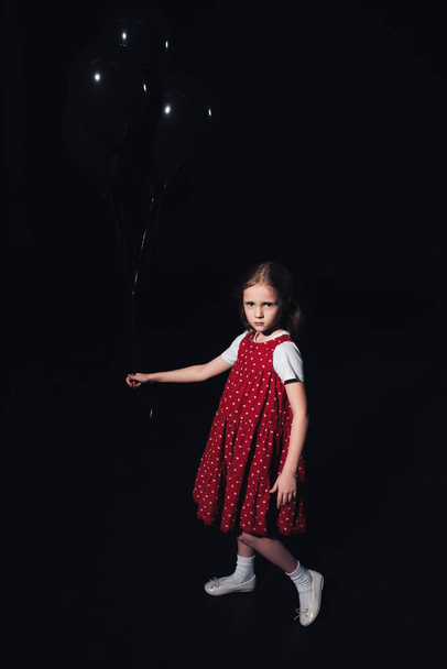 offended kid holding black balloons and looking at camera isolated on black - Zdjęcie, obraz