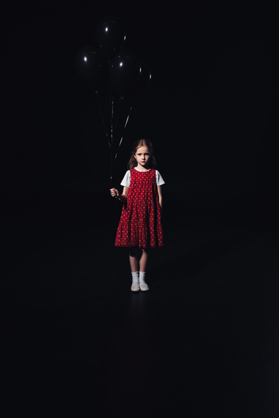 smutné dítě, které stojí s černými balónky a dívá se na fotoaparát izolovaný na černém - Fotografie, Obrázek