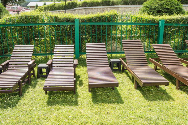 As cadeiras de madeira do convés estão em uma fileira na grama verde sob o sol na área de recreação no território privado de um hotel de luxo moderno. Jardim para banhos de sol e relaxamento
 - Foto, Imagem