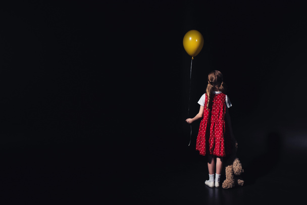 back view of depressed child with yellow balloon and teddy bear isolated on black - Photo, Image