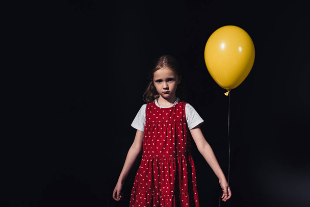 offended, lonely child with yellow balloon looking at camera isolated on black - Foto, afbeelding