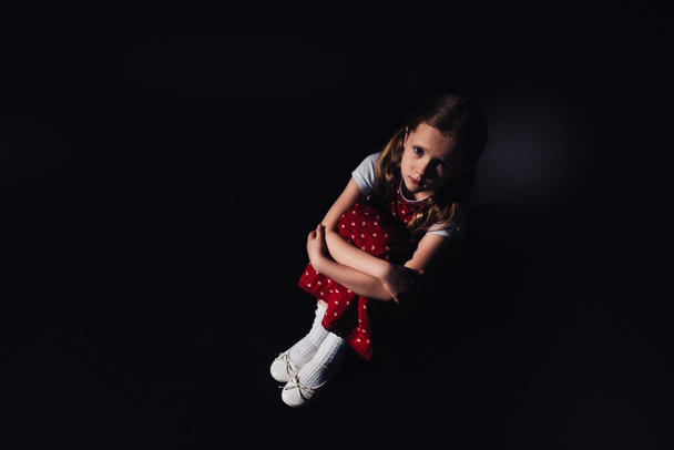 грустный, испуганный ребенок сидит на полу и смотрит в камеру на черном фоне
 - Фото, изображение