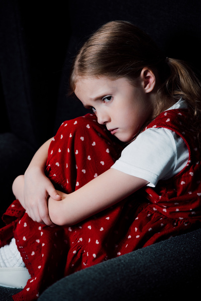 offended, frightened kid sitting in armchair isolated on black - Photo, Image