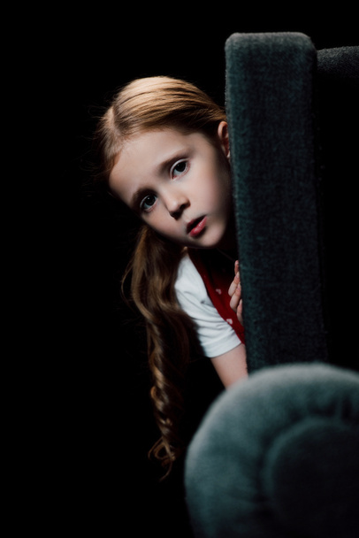 anxious child looking at camera while hiding behind armchair isolated on black - Photo, Image