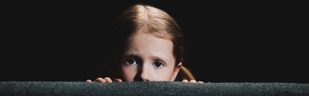 panoramic shot of frightened child hiding behind armchair and looking at camera  isolated on black - Zdjęcie, obraz