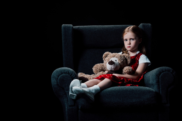 trauriges, einsames Kind im Sessel sitzend und Teddybär isoliert auf schwarz haltend - Foto, Bild
