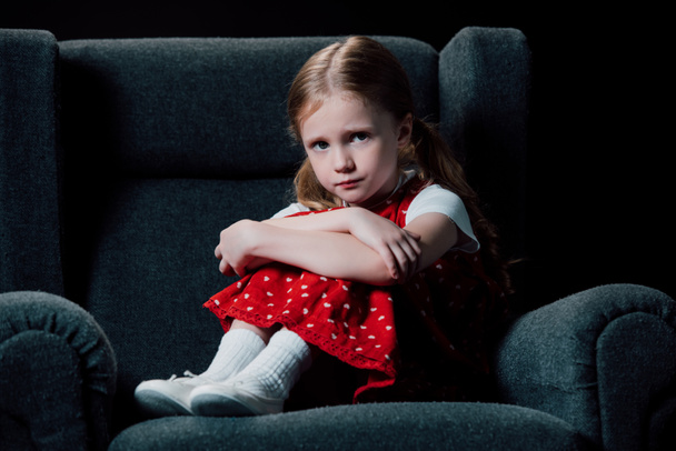 deprimovaný, vystrašený dítě sedící v křesle a pohled na fotoaparát izolovaný na černém - Fotografie, Obrázek