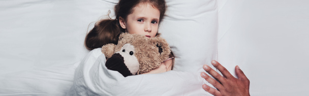 partial view of male hand near scared child lying in bed with teddy bear, panoramic shot - Fotó, kép