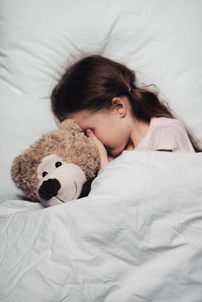 entzückendes Kind, das Augen mit der Hand bedeckt, während es mit Teddybär im Bett liegt - Foto, Bild