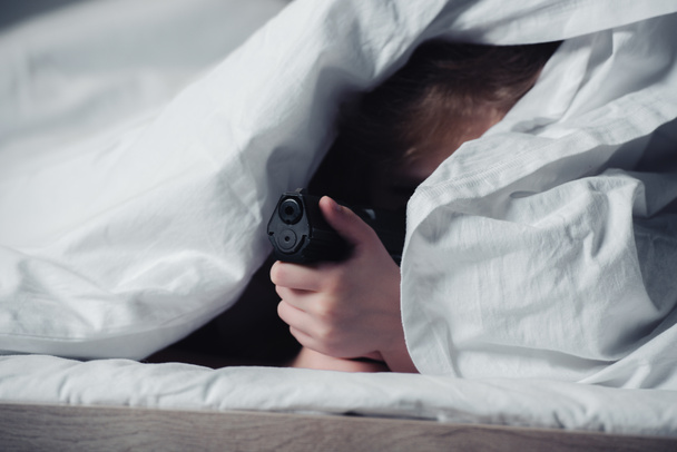 scared child holding gun while hiding under blanket in bedroom - Фото, изображение