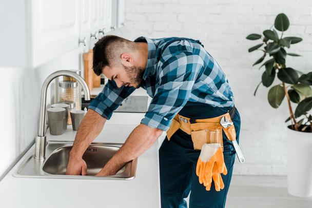 handsome bearded repairman working near sink in kitchen  - Фото, изображение
