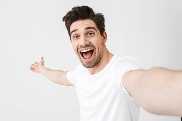 Podekscytowany szczęśliwy młody człowiek pozowanie na białym tle ściany zrobić selfie przez aparat fotograficzny. - Zdjęcie, obraz