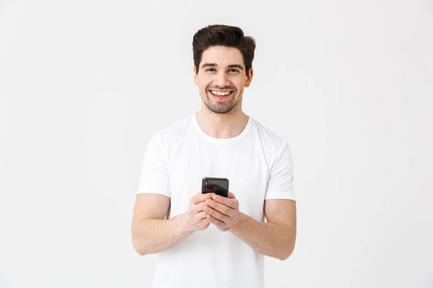 Захоплений щасливий молодий чоловік позує ізольовано на білому тлі стіни за допомогою мобільного телефону
. - Фото, зображення