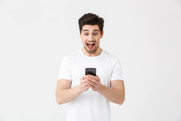 Excited happy young man posing isolated over white wall background using mobile phone. - 写真・画像