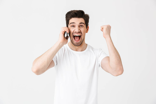 Boldog izgatott döbbent fiatalember pózol elszigetelt fehér fal háttérben beszél a mobil telefon. - Fotó, kép