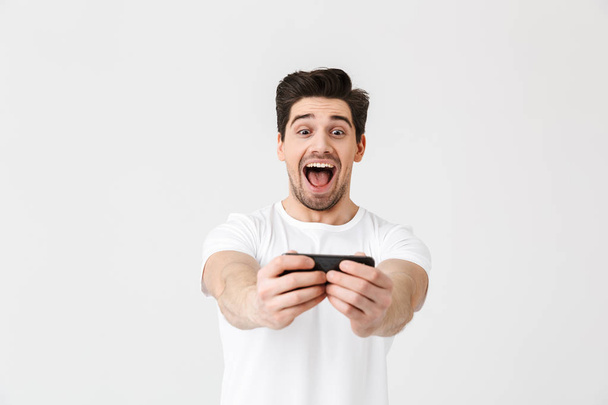 Innoissaan onnellinen emotionaalinen nuori mies poseeraa yli valkoinen seinä tausta pelata pelejä matkapuhelimella
. - Valokuva, kuva