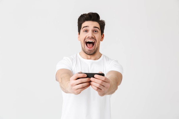 Excited happy emotional young man posing isolated over white wall background play games by mobile phone. - Valokuva, kuva