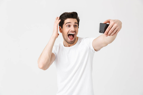 Happy emotional young man posing isolated over white wall background take selfie by mobile phone. - Фото, зображення