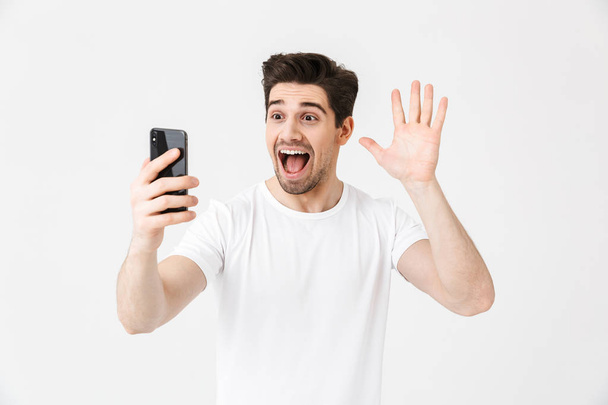 Young man posing isolated over white wall background using mobile phone take a selfie. - Фото, изображение