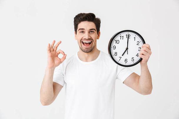 Shocked excited happy young man posing isolated over white wall background holding clock. - Fotoğraf, Görsel