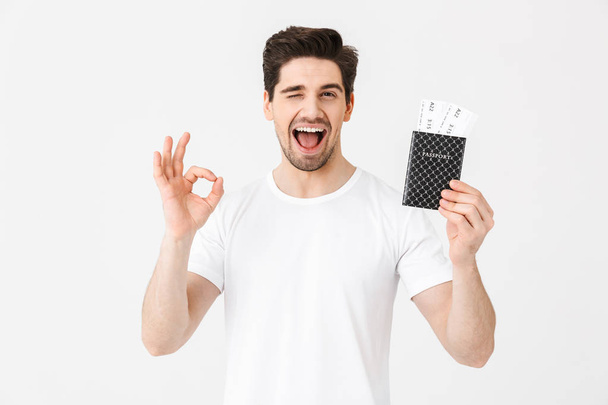 Happy young man posing isolated over white wall background holding passport with tickets showing okay gesture. - Foto, immagini