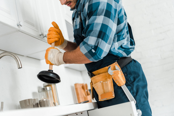 cropped view of handyman with tool belt holding plunger in kitchen  - Photo, Image