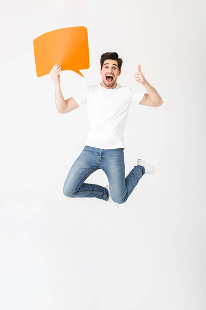 Emotional excited young man posing isolated over white wall background holding speech bubble. - Фото, изображение