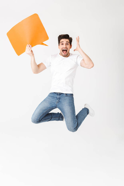 Emotional excited young man posing isolated over white wall background holding speech bubble. - Foto, afbeelding