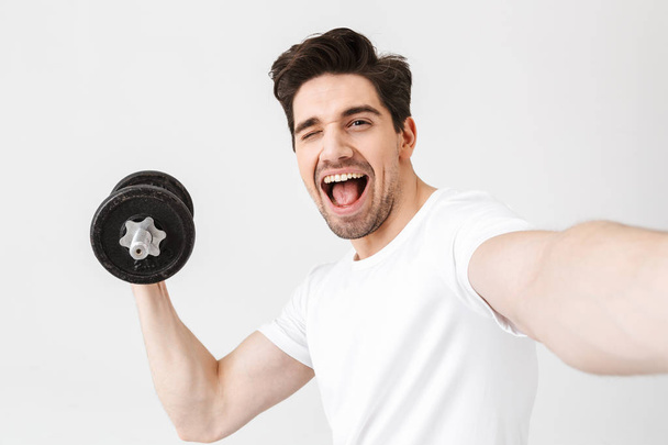 Emotional happy excited young man posing isolated over white wall holding dumbbell make exercise take a selfie by camera. - Zdjęcie, obraz