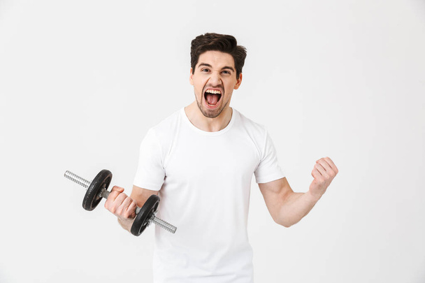 Screaming excited young man posing isolated over white wall holding dumbbell make exercise. - Φωτογραφία, εικόνα