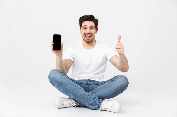 Excited young man posing isolated over white wall background using mobile phone showing display sitting on floor. - 写真・画像
