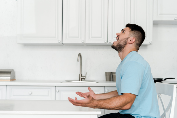 side view of upset man screaming while sitting near table in kitchen  - Photo, Image