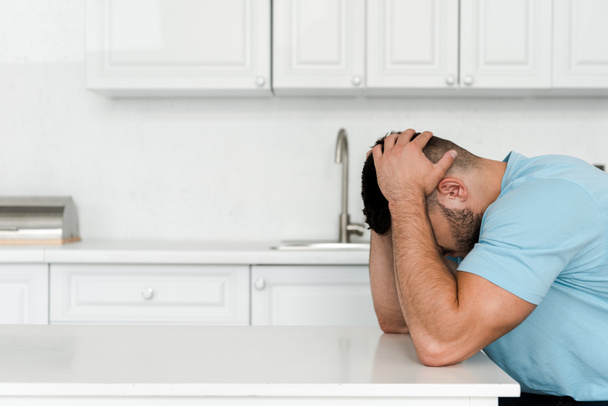 frustrado hombre tocando la cabeza mientras está sentado cerca de la mesa en la cocina
  - Foto, Imagen