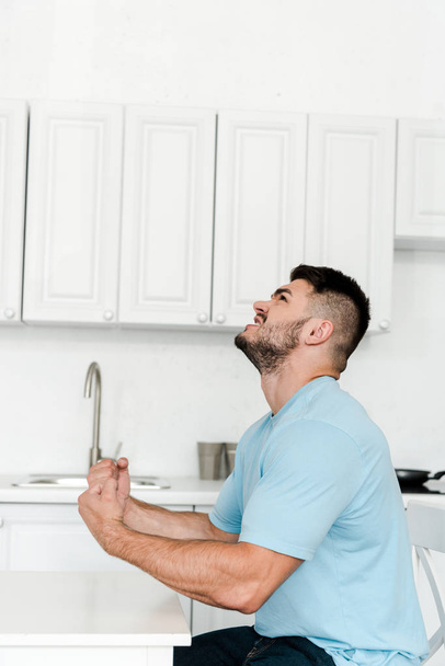 Вид сбоку сердитого мужчины, кричащего, сидя за столом на кухне
  - Фото, изображение