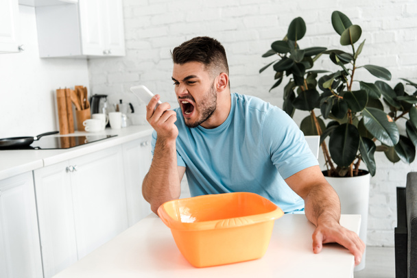 selective focus of upset bearded man screaming at smartphone near plastic wash bowl  - Photo, Image