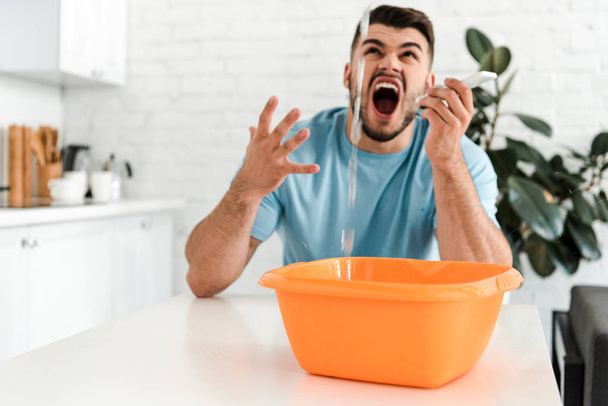 selective focus of bearded man screaming while looking at water pouring into plastic wash bowl  - Foto, Imagen