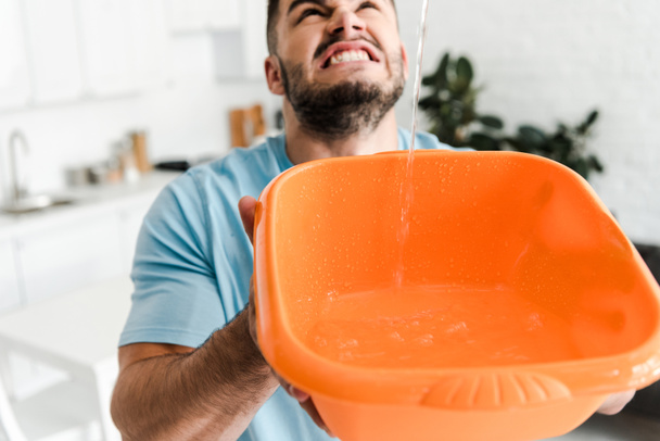 selective focus of displeased bearded man holding plastic wash bowl near pouring water  - Foto, immagini