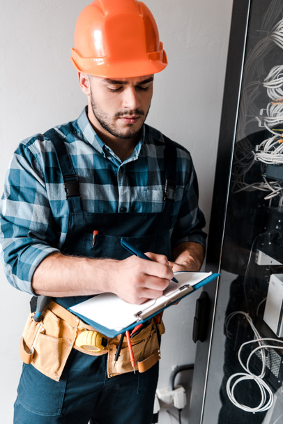 handsome technician holding clipboard while writing near wires and cables  - Photo, Image