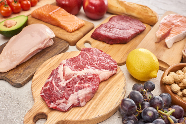 Fresh raw meat, poultry, fish on wooden cutting boards near lemon, grapes, apples, branch of cherry tomatoes, nuts and french baguette on marble surface - Photo, Image