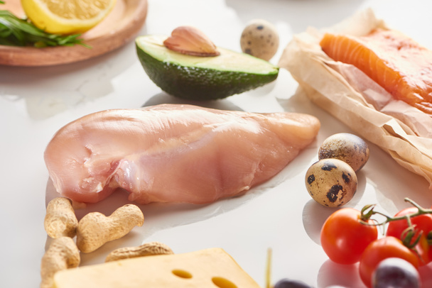 Κλείσιμο της προβολής των ακατέργαστων πουλερικών και φιλέτο σολομού κοντά σε αυγά ορτυκιού, φιστίκια, ντομάτες, αβοκάντο, λεμόνι και τυρί σε λευκή επιφάνεια - Φωτογραφία, εικόνα