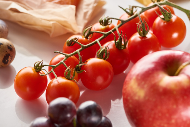 Close up view of branch of cherry tomatoes near apple, grape and quail eggs on white surface - Foto, afbeelding