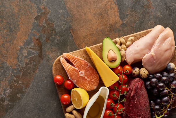 Ansicht des Holzschneidebretts mit rohem Fisch, Fleisch, Geflügel, Käse, Obst, Gemüse, Olivenöl und Erdnüssen auf dunkelbrauner Marmoroberfläche mit Kopierraum - Foto, Bild