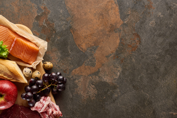 Vue de dessus de la planche à découper en bois avec poisson cru, bacon, raisin, pomme, baguette et oeufs de caille sur une surface en marbre brun foncé avec espace de copie
 - Photo, image