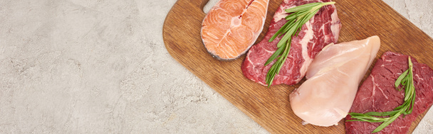 Panoramic shot of raw meat, fish and poultry with rosemary twigs on wooden cutting board on marble surface with copy space - Photo, Image